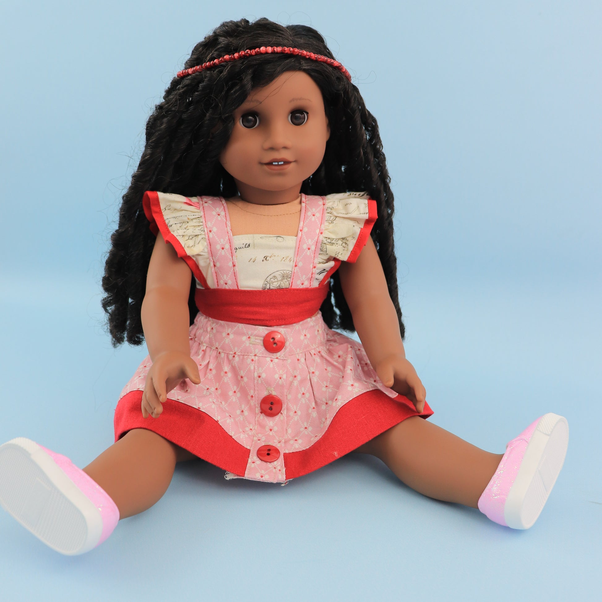 American girl doll, dressed in a flutter sleeve top and skirt. The skirt has a hem trim and is buttoned down the front. Both top and skirt are made using Michael Miller fabrics in pink cotton with a geometric pattern and a Letter themed print.