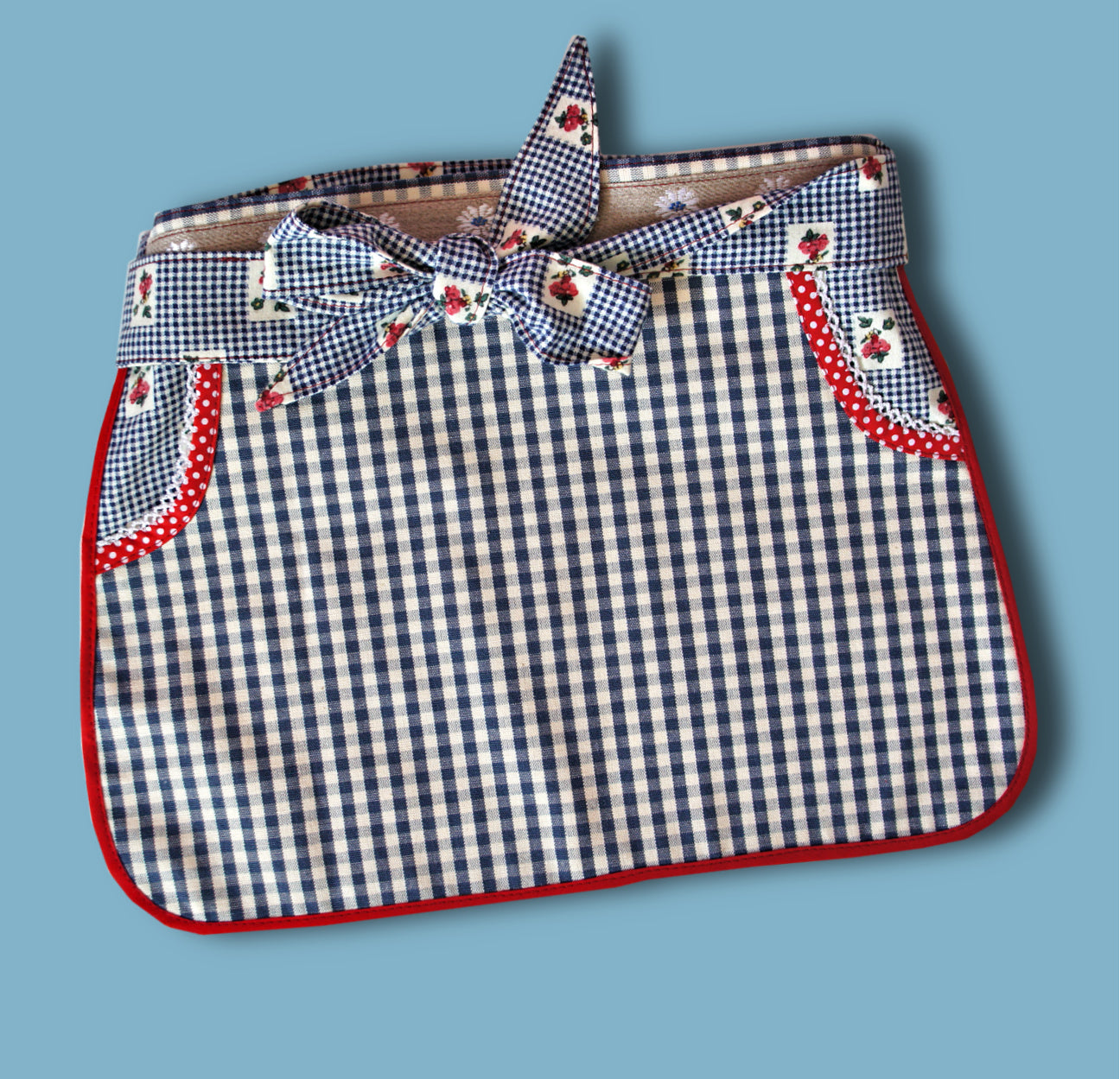 Half apron Pdf sewing pattern by Frocks & Frolics. Fully lined, cute tie backs, free video sew-a-long. In gingham white and blue cotton with red trim bias binding and pockets.