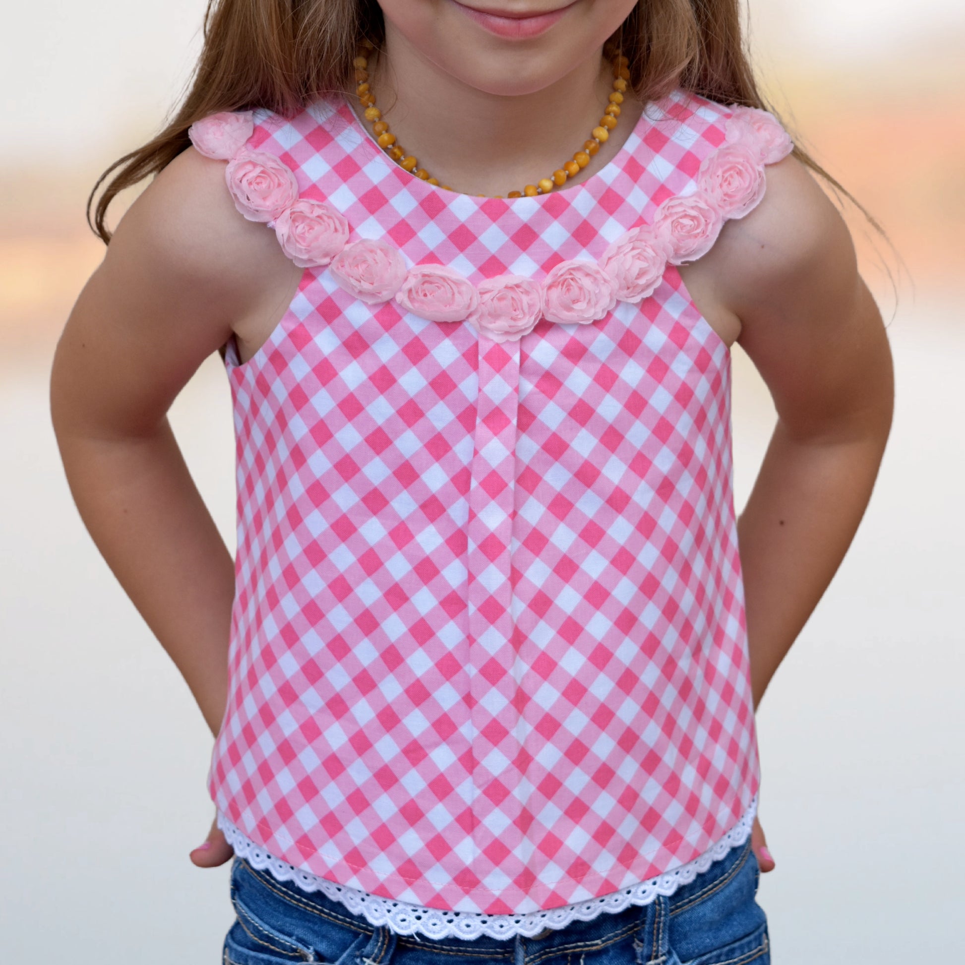 Pretty girl wearing a cute blouse with round yoke collar. Its in a Michael Miller Gingham and has decorative roses attached to the round yoke. The side view reveals a fully lined armhole. The hem has a slim lace trim.