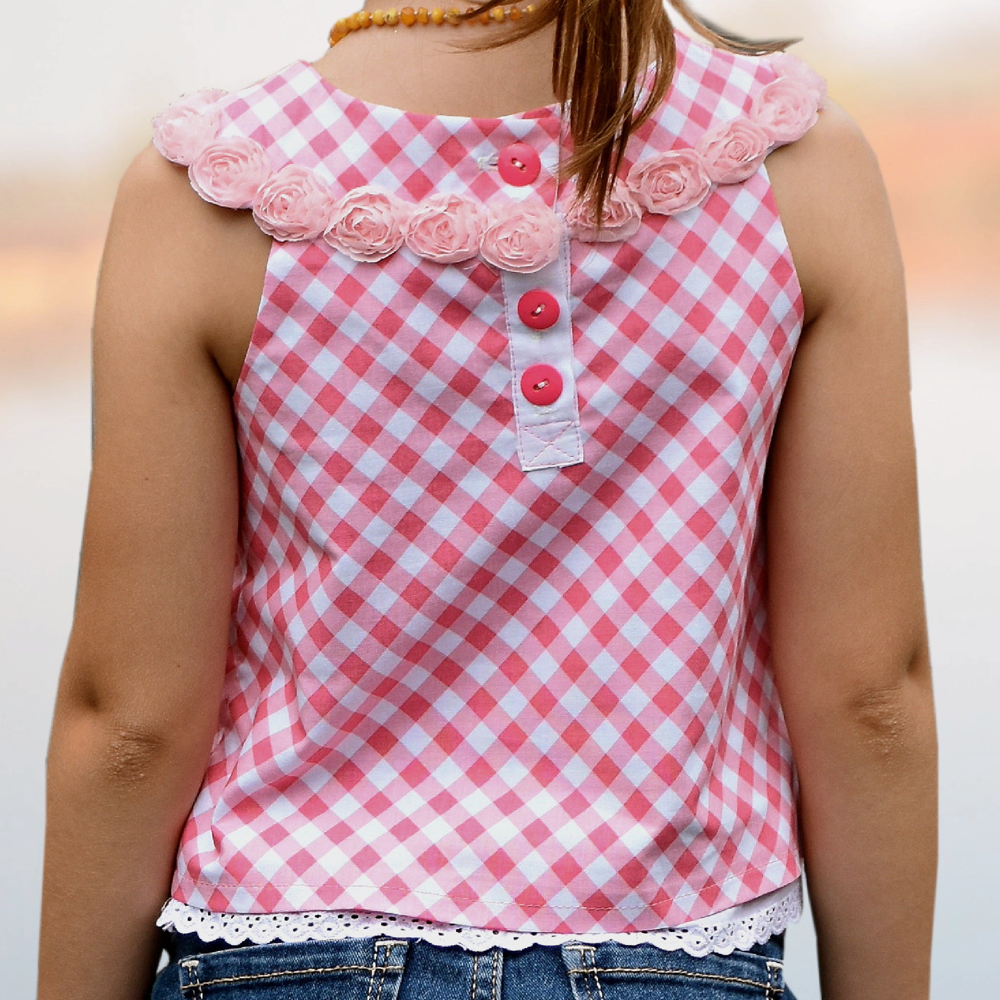 Pretty girl wearing a cute blouse with round yoke collar. Its in a Michael Miller Gingham and has decorative roses attached to the round yoke. The side view reveals a fully lined armhole. The hem has a slim lace trim. Plkacket closure.