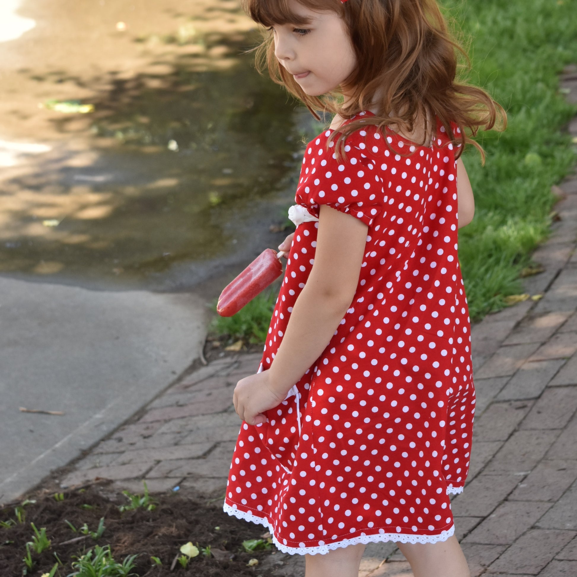 Millie dress pdf sewing pattern by Frocks & Frolics. Empire line bodice and slightly gathered skirt. Puff sleeves with elastication. Patch pockets with piping. Bodice has a cute lace trim. The fabric is white polka dots on a red cotton.