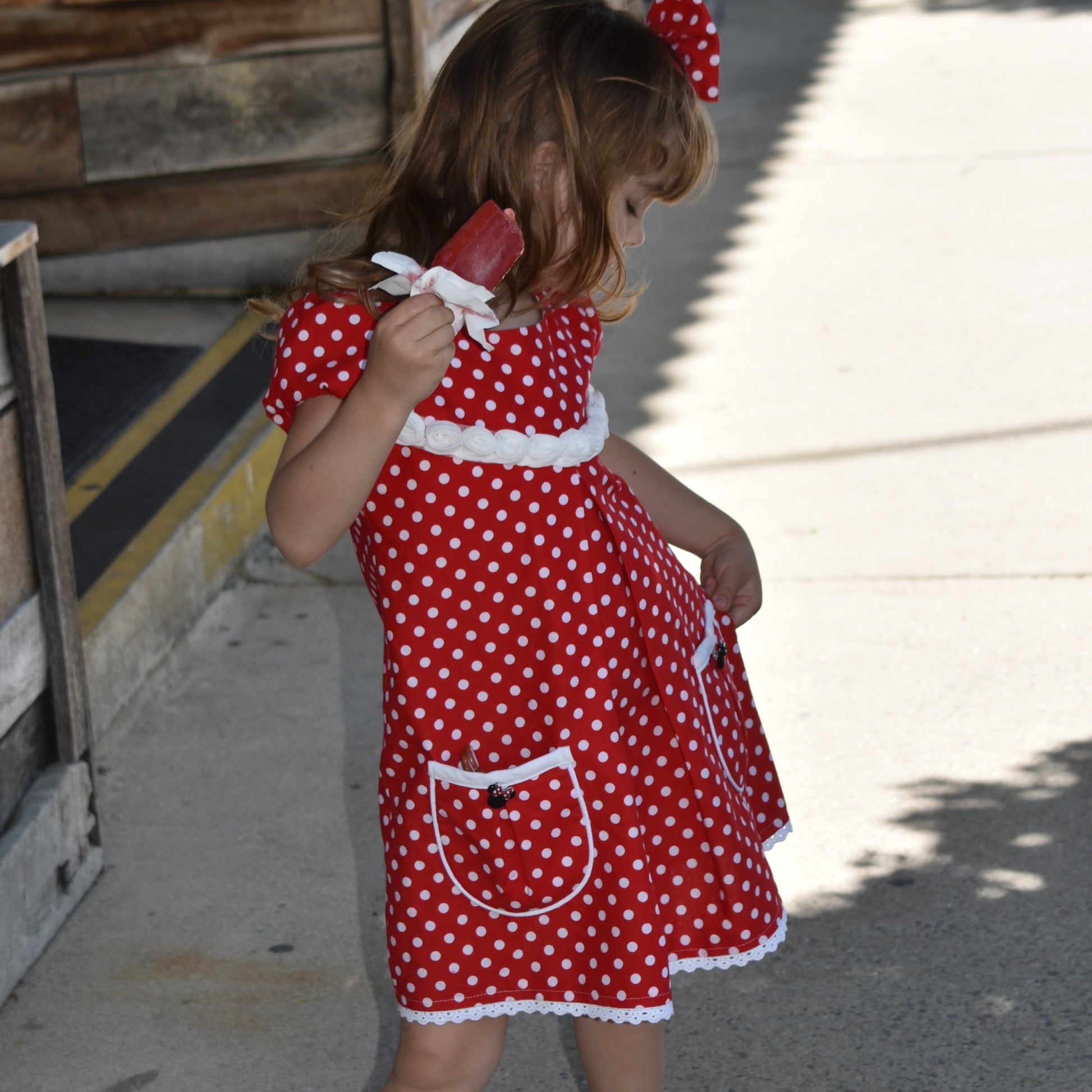 Millie dress pdf sewing pattern by Frocks & Frolics. Empire line bodice and slightly gathered skirt. Puff sleeves with elastication. Patch pockets with piping. Bodice has a cute lace trim. The fabric is white polka dots on a red cotton.