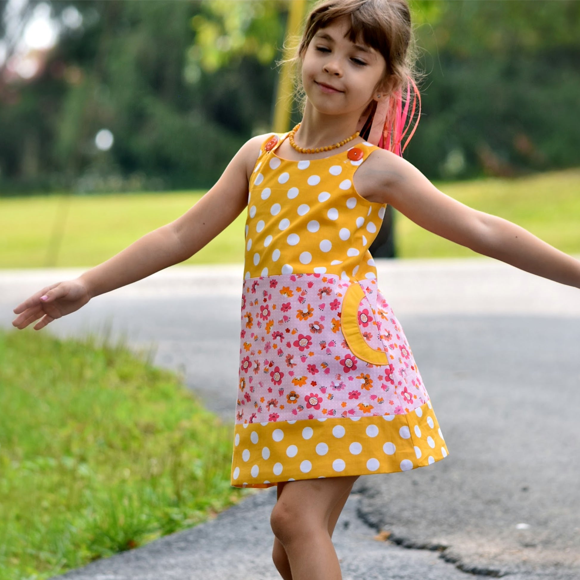 Mimi apron dress pdf sewing pattern. For girls age 1-12. Fully lined with side pockets and cute applique. In white polka dots with mint back base. Colour blocking with a flower print. Front and back view.