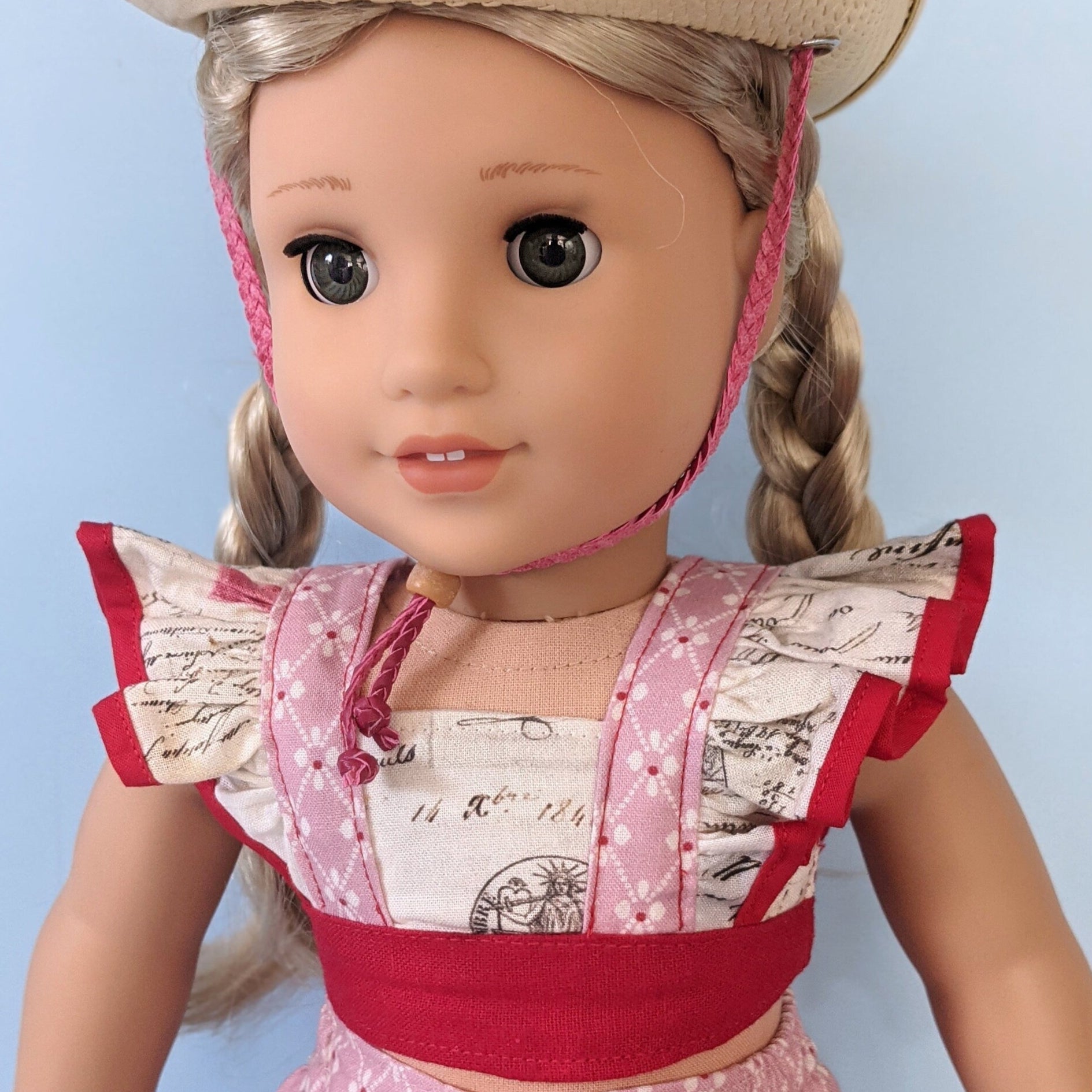 American girl doll, dressed in a flutter sleeve top and skirt. The skirt has a hem trim and is buttoned down the front. Both top and skirt are made using Michael Miller fabrics in pink cotton with a geometric pattern and a Letter themed print.