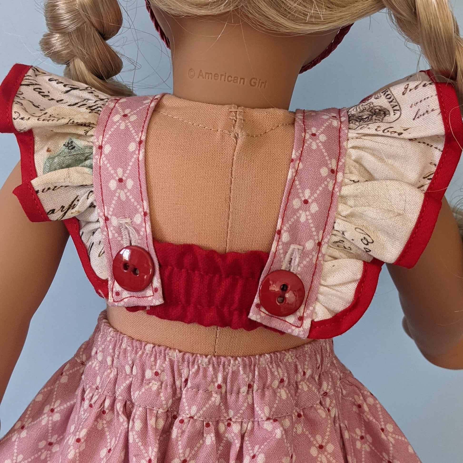 American girl doll, dressed in a flutter sleeve top and skirt. The skirt has a hem trim and is buttoned down the front. Both top and skirt are made using Michael Miller fabrics in pink cotton with a geometric pattern and a Letter themed print.
