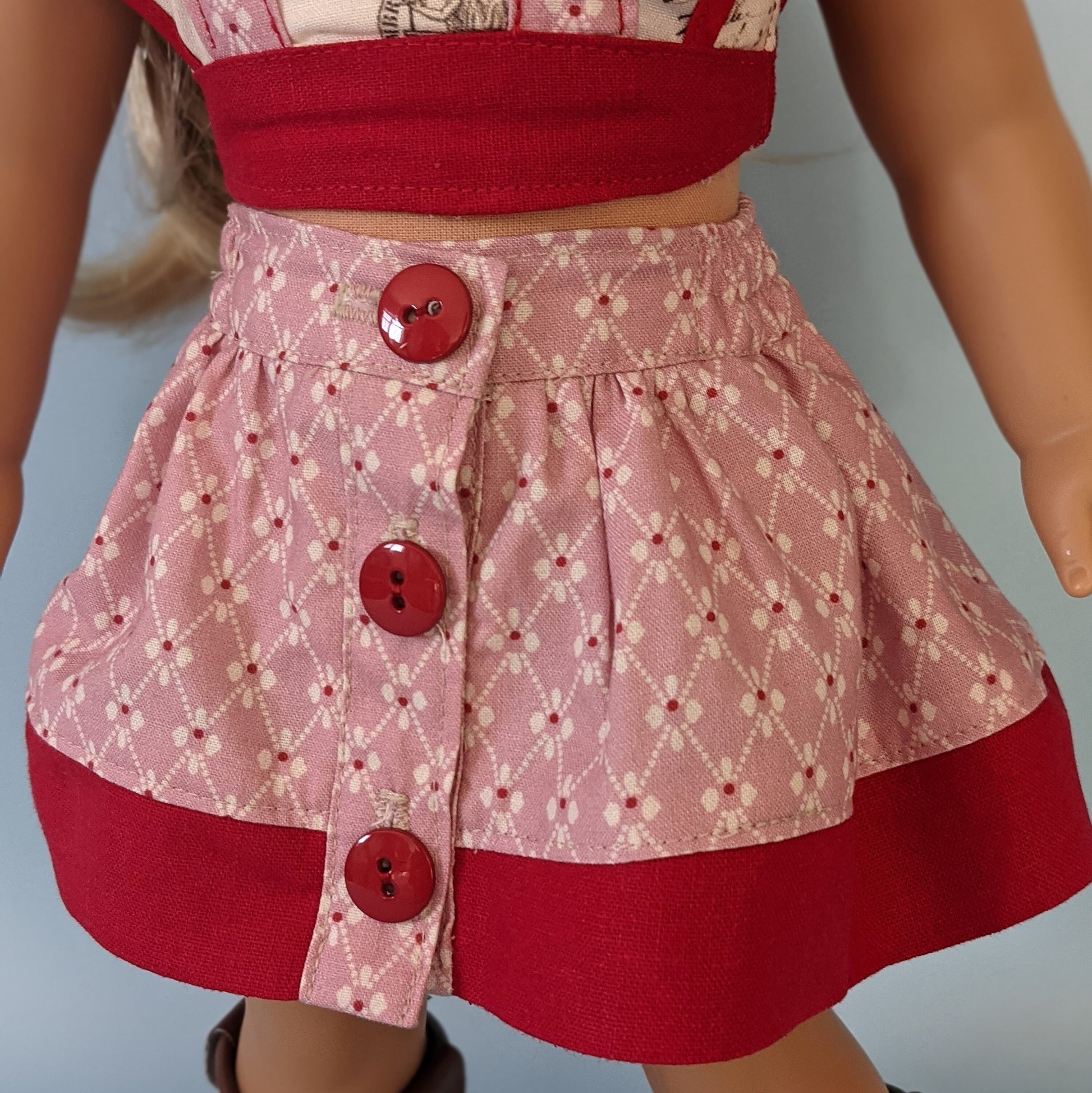 American girl doll, dressed in a flutter sleeve top and skirt. The skirt has a hem trim and is buttoned down the front. Both top and skirt are made using Michael Miller fabrics in pink cotton with a geometric pattern and a Letter themed print.