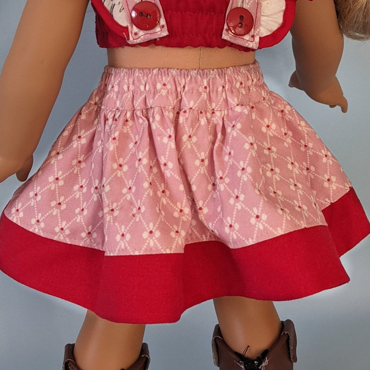 American girl doll, dressed in a flutter sleeve top and skirt. The skirt has a hem trim and is buttoned down the front. Both top and skirt are made using Michael Miller fabrics in pink cotton with a geometric pattern and a Letter themed print.