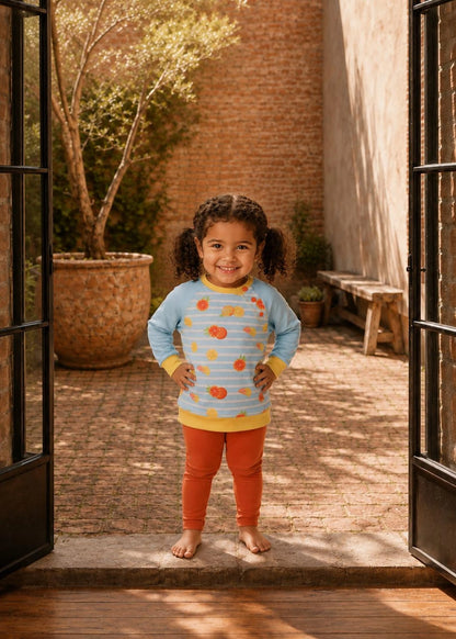 A young child stands barefoot in a doorway of a sunlit courtyard, wearing a light blue long-sleeve top with a colorful fruit print, yellow trim, and orange pants.