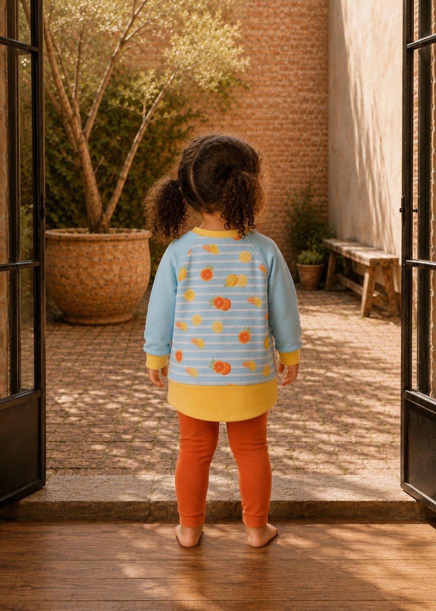 A toddler viewed from behind wearing a light blue striped long-sleeve tunic top with a yellow hem and orange fruit print, paired with orange leggings, standing in an open doorway looking into a sunlit courtyard.