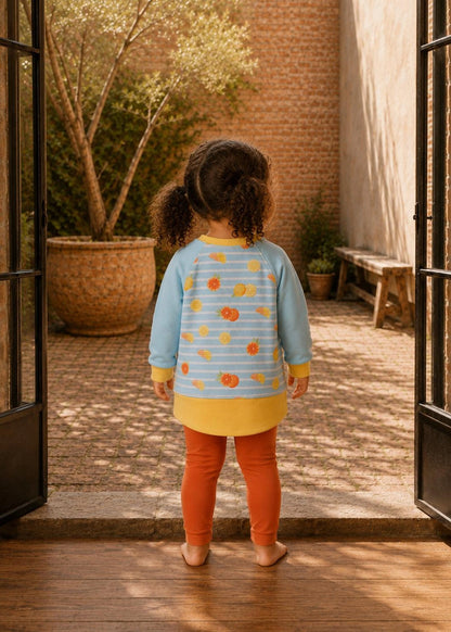 A toddler viewed from behind wearing a light blue striped long-sleeve tunic top with a yellow hem and orange fruit print, paired with orange leggings, standing in an open doorway looking into a sunlit courtyard.
