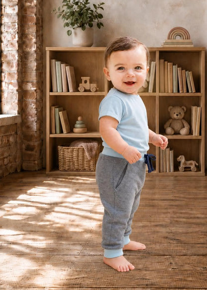 A toddler standing barefoot in a sunlit room, wearing a light blue short-sleeve t-shirt and heather gray pants with a small blue bow tie detail at the waist. The background features a wooden bookshelf with toys and books.