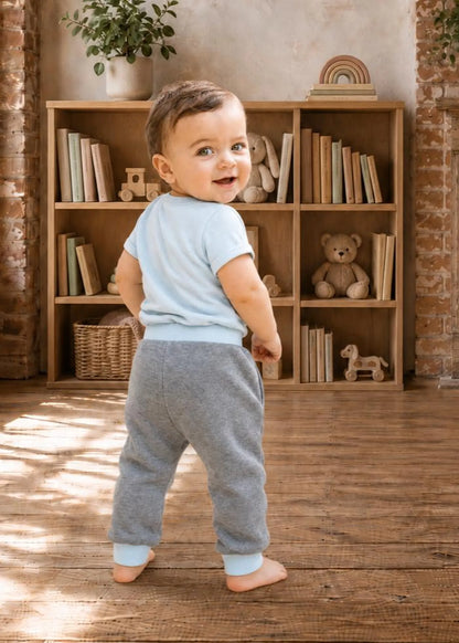 A toddler standing barefoot in a room with wooden floors and a bookshelf, wearing a light blue short-sleeve t-shirt and heather gray jogger pants.