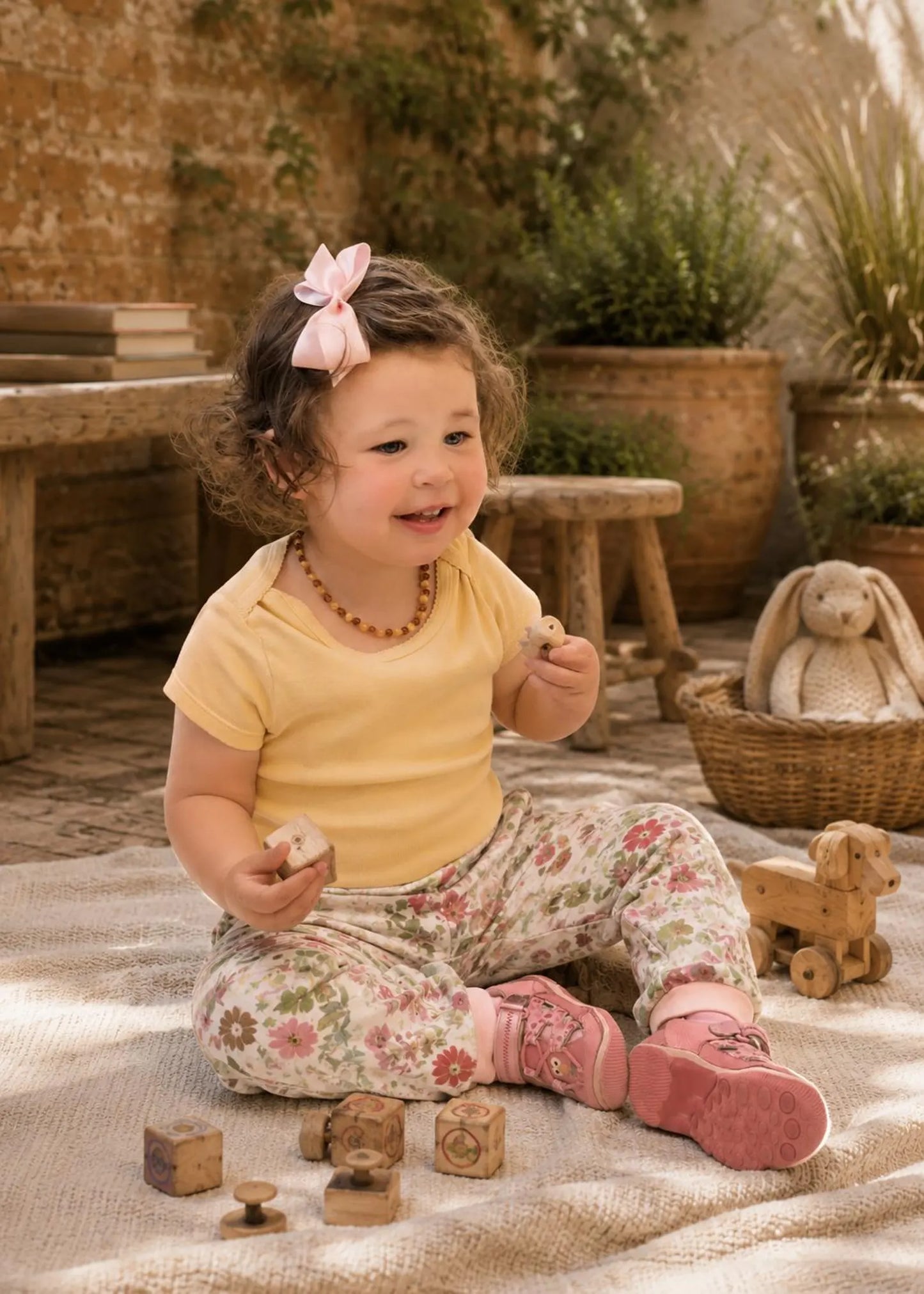 A toddler girl sitting on a beige blanket in a rustic playroom, wearing a pale yellow short-sleeve t-shirt and floral print leggings with pink sneakers. She has a pink bow in her hair and is holding a wooden block.