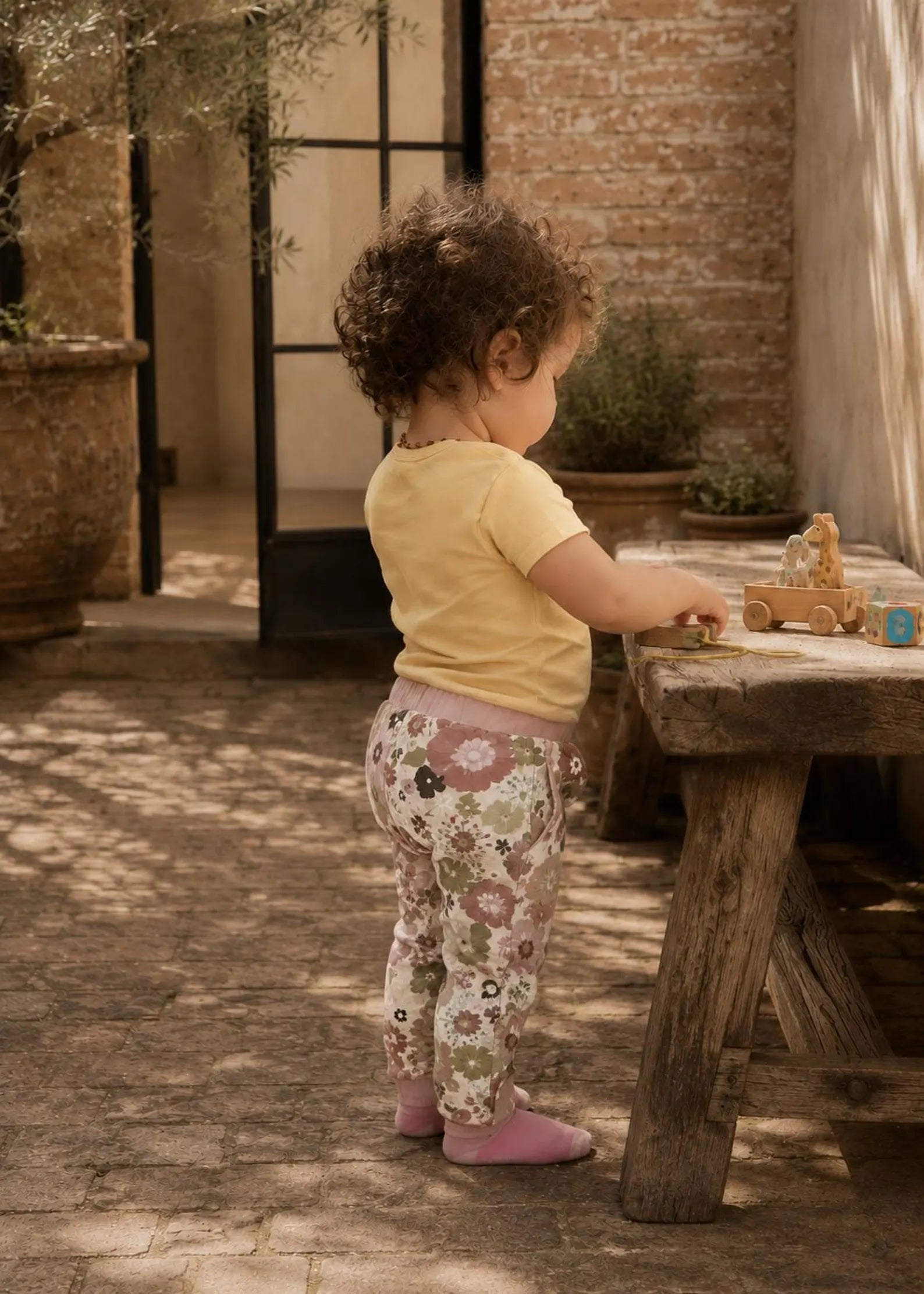 A toddler stands at a rustic wooden table in a sunlit courtyard, wearing a pale yellow short-sleeve top and floral print leggings with pink socks. A small wooden toy is on the table.