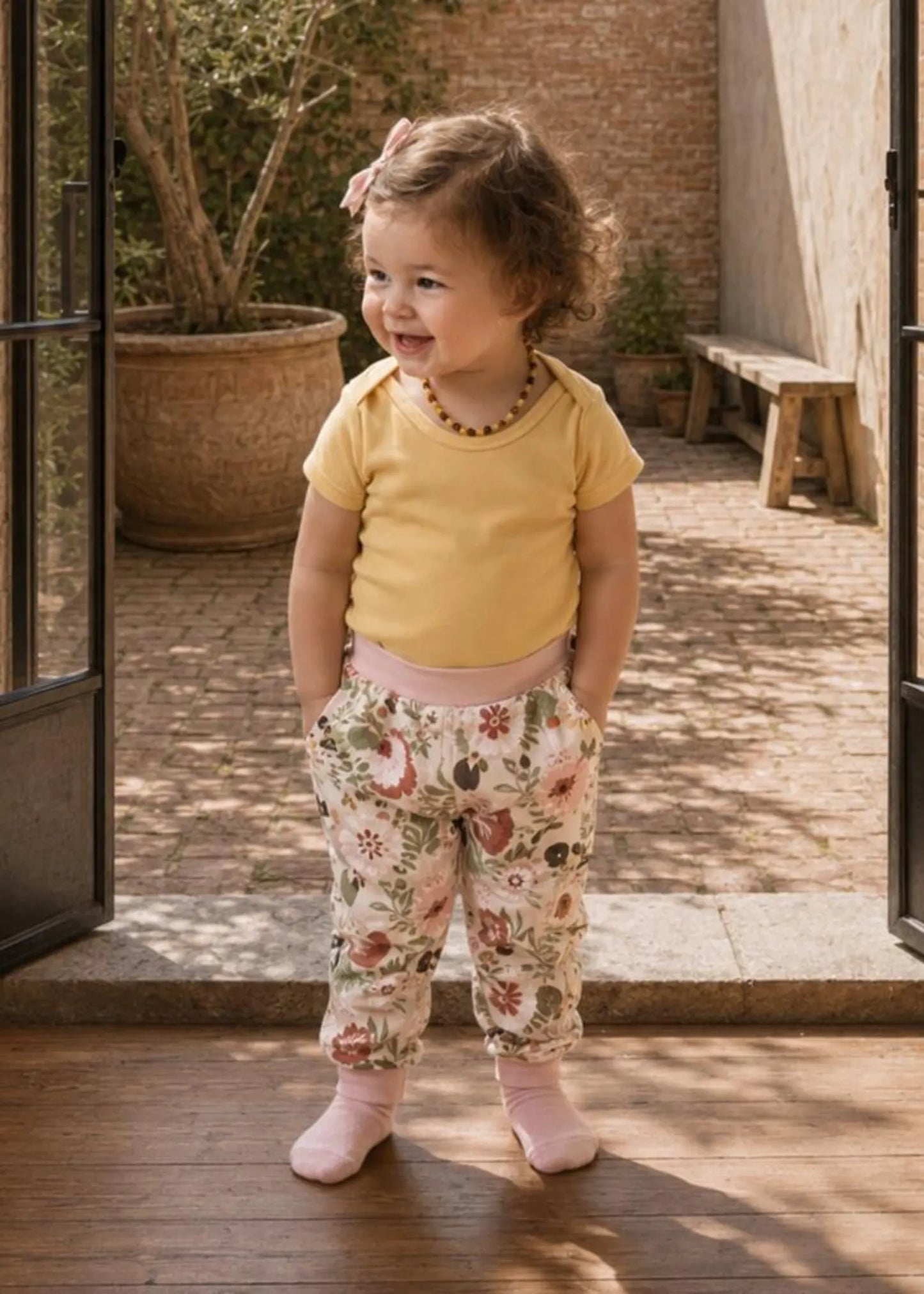 A toddler girl stands in a sunlit courtyard doorway wearing a pale yellow short-sleeve top, floral print pants, and pink socks.