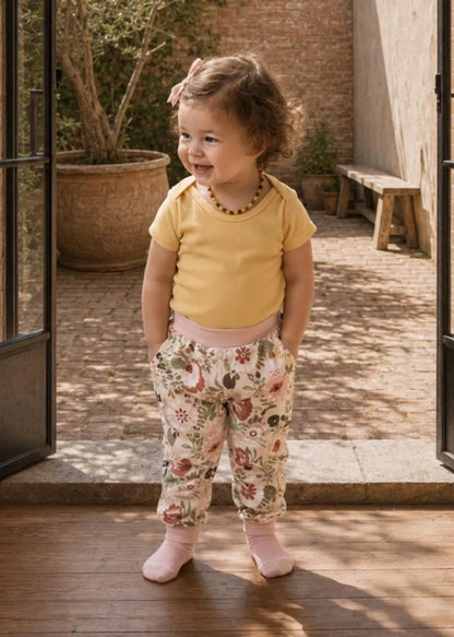 A toddler girl stands in a sunlit courtyard doorway wearing a pale yellow short-sleeve top, floral print pants, and pink socks.