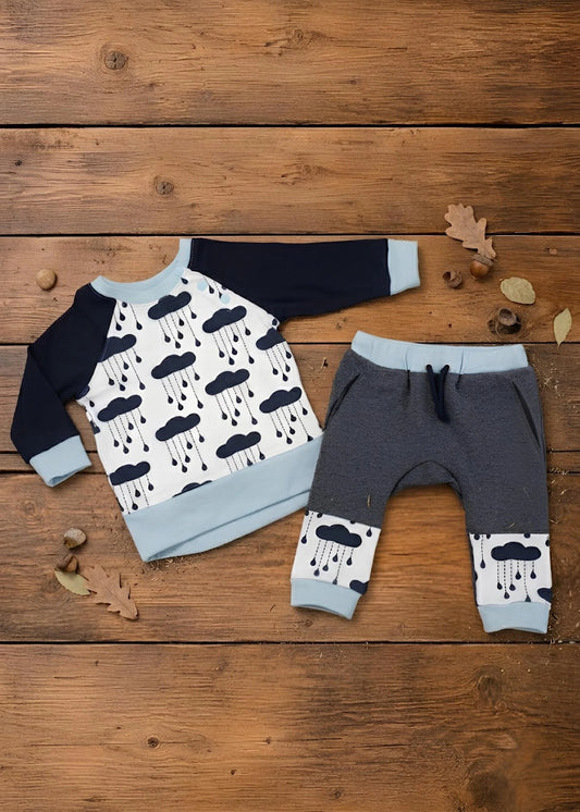 A baby clothing set laid flat on a rustic wooden surface, featuring a long-sleeve raglan sweatshirt and matching pants. The sweatshirt has a white body with a black cloud and raindrop print, navy sleeves, and light blue cuffs and hem.