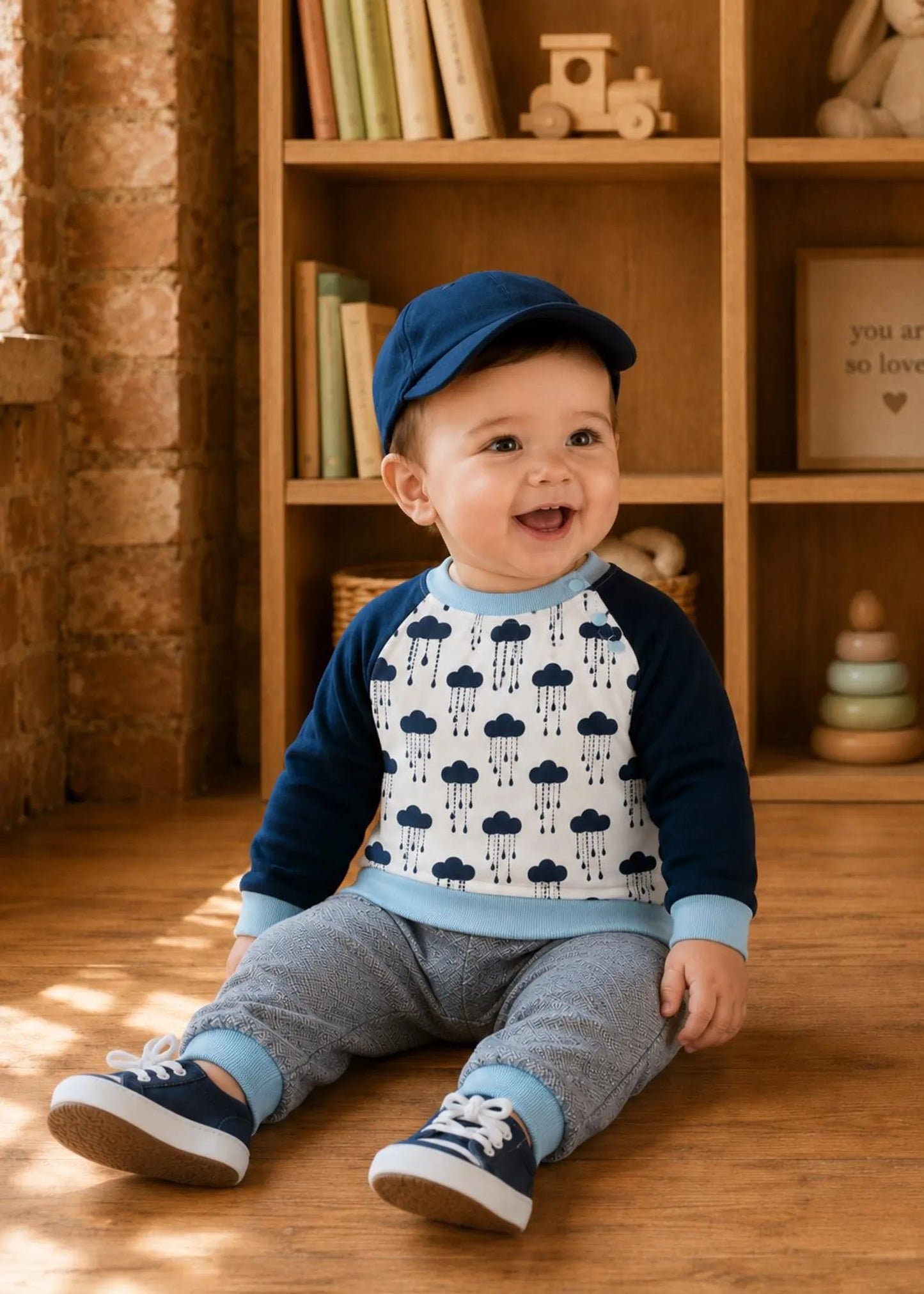 A baby sitting on a wooden floor in front of a bookshelf, wearing a navy blue and light blue raglan long-sleeve top with a dark blue raincloud pattern on the front, gray pants, and a navy blue cap.