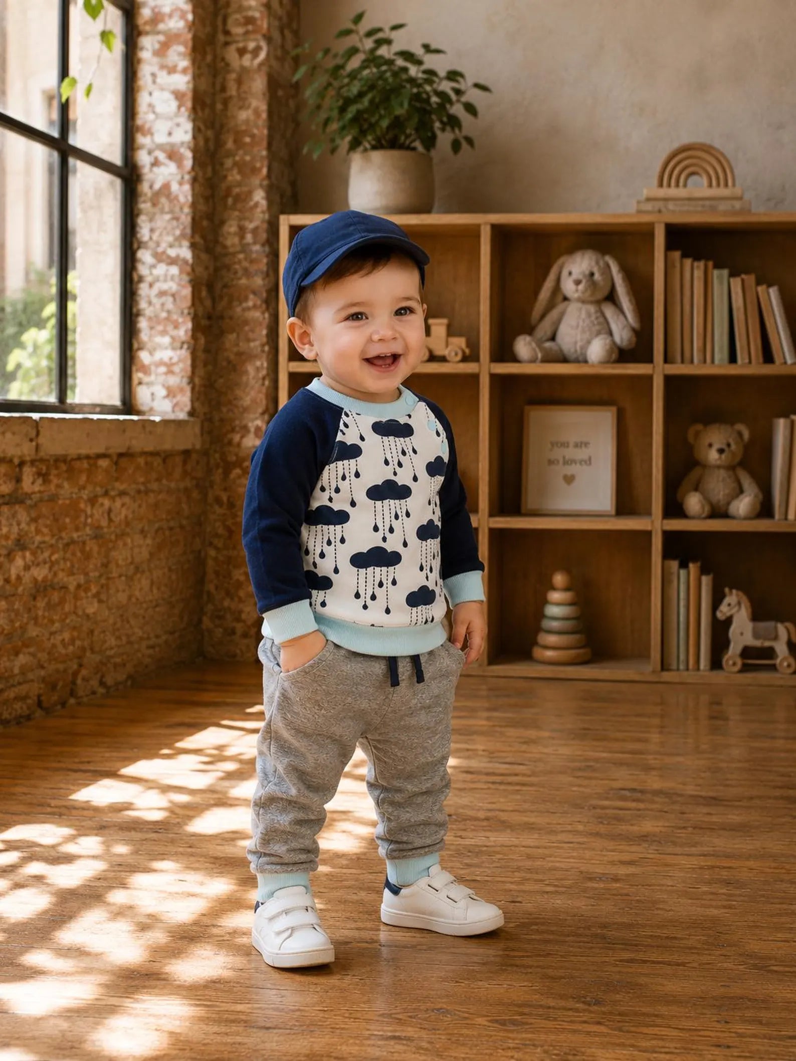 A baby boy wearing a navy blue and white raglan long-sleeve shirt with a cloud pattern, gray joggers, and white sneakers, standing in a sunlit room with a wooden bookshelf in the background.