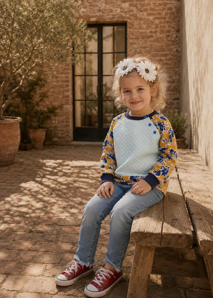 A young girl with blonde curly hair wearing a white flower headband sits on a rustic wooden bench in an outdoor courtyard. She is wearing a raglan-style long-sleeve top with a light blue textured body, navy blue collar and cuffs, and yellow sleeves with a blue floral pattern, paired with blue jeans and red sneakers.
