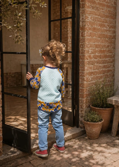 A young child viewed from behind wearing a light blue textured long-sleeve top with a yellow and blue patterned hem and cuffs, paired with light blue jeans and red sneakers, standing at an open black-framed glass door in a rustic brick courtyard.