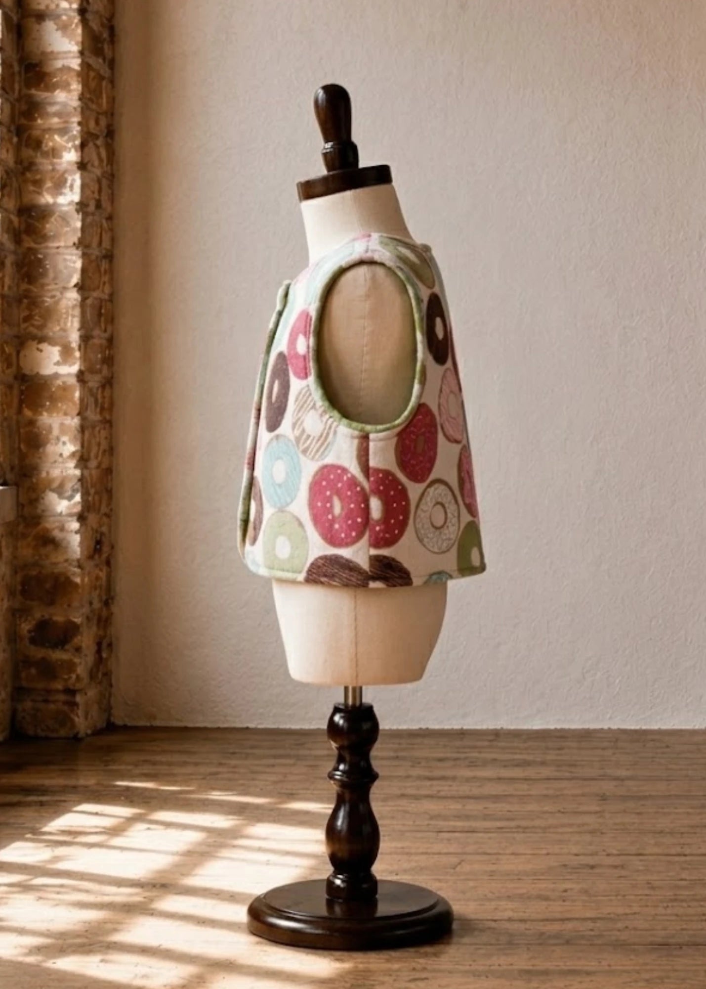 A vest with donut print, displayed on a cream mannequin torso mounted on a dark wooden stand in a sunlit room.