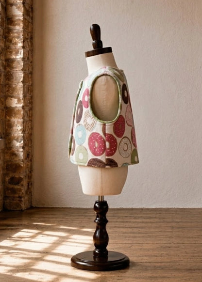 A vest with donut print, displayed on a cream mannequin torso mounted on a dark wooden stand in a sunlit room.