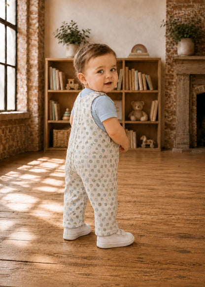 A toddler standing side view in a sunlit room with exposed brick walls and wooden floors, wearing light blue short-sleeve top and cream overall with small blue polca dot print.