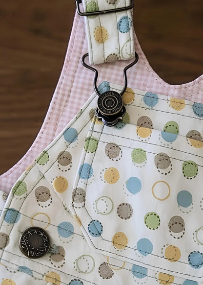 Close-up view of handmade overalls featuring a colorful polka dot fabric, a pink gingham inner lining, and brass hardware including an overall clasp and engraved side button.