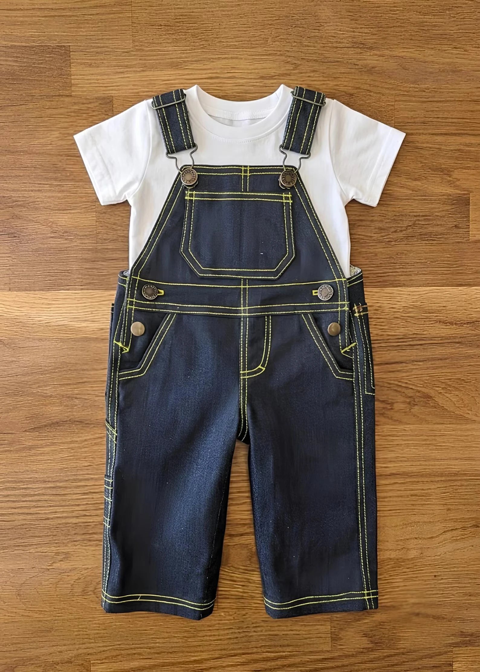 Dark indigo denim toddler overalls featuring bright yellow contrast stitching and metal hardware, layered over a classic white short-sleeve t-shirt.