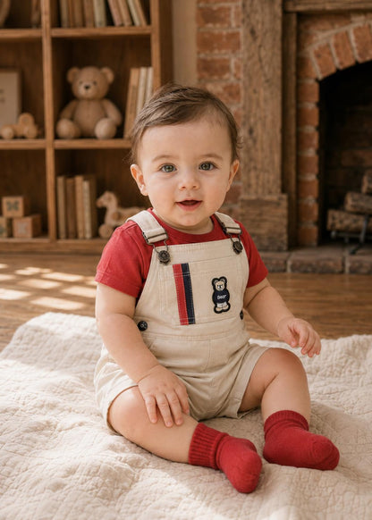 A toddler sitting on a cream blanket in a cozy room, wearing beige overalls over a red t-shirt and red socks. The overalls feature a small embroidered bear on the chest with a red and navy stripe detail.