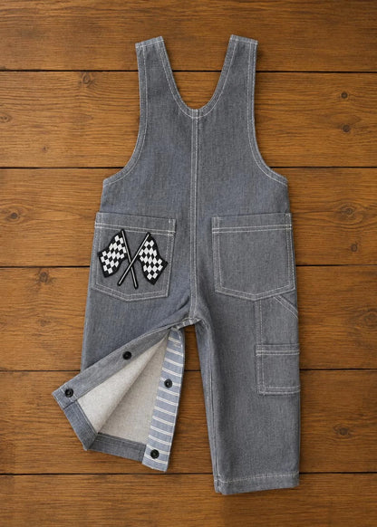 Grey denim toddlers' overalls laid flat on a wooden surface, featuring crossed checkered racing flags embroidered on the left back pocket and snap-closure inner legs.