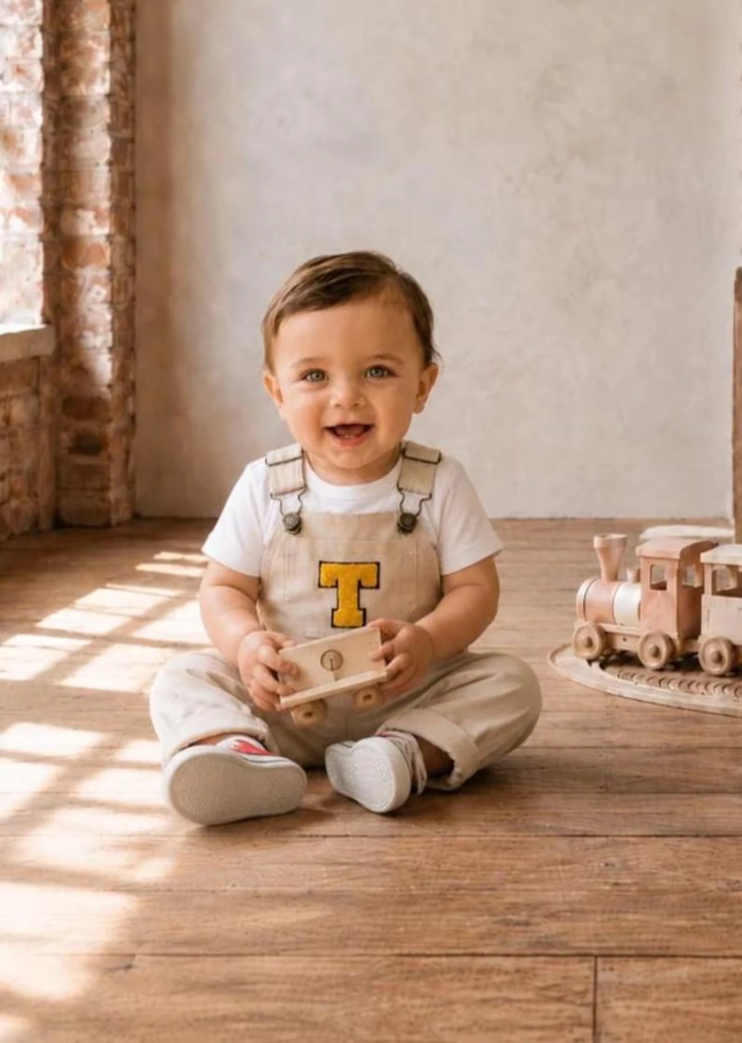 Smiling baby boy sitting on a wooden floor in light beige overalls featuring a varsity-style yellow letter "T" patch on the chest, layered over a white t-shirt.
