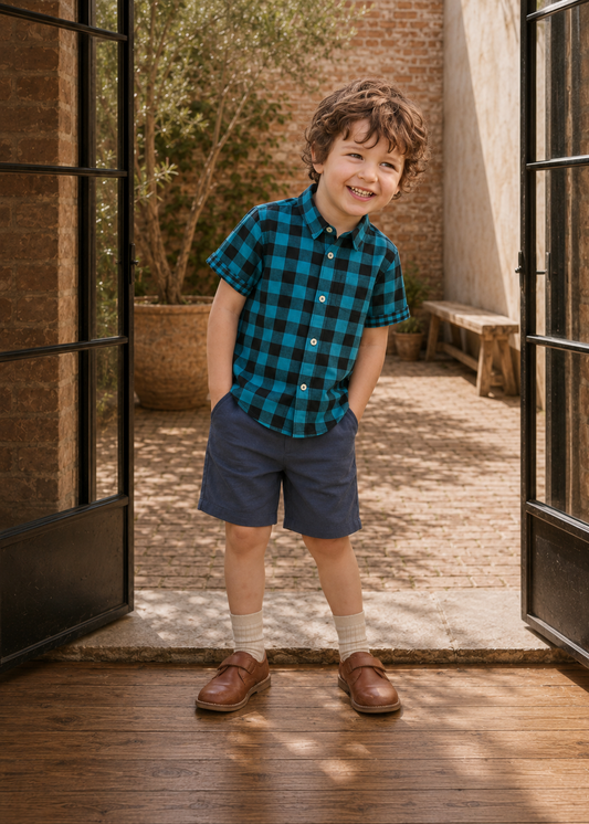 Front view of a boy in a teal and black plaid Cool Shirt and navy shorts standing by the doorway