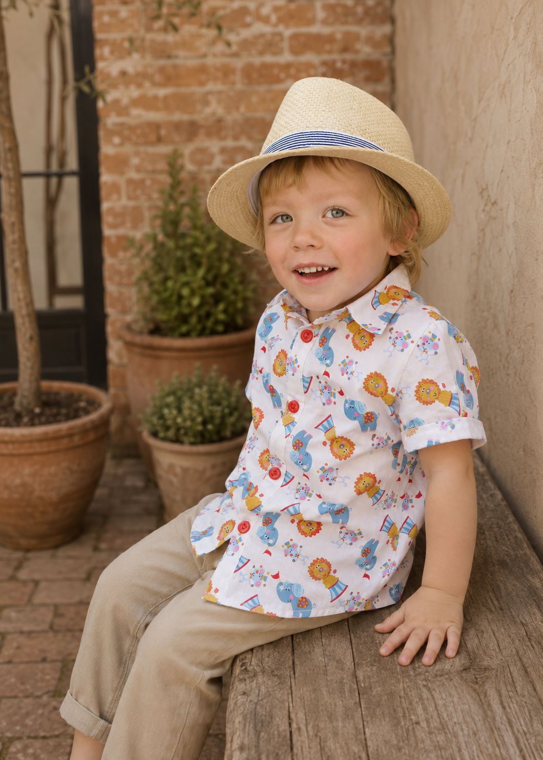 Toddler in a straw hat sitting on a wooden bench wearing a circus animal print Cool Shirt with tan pants