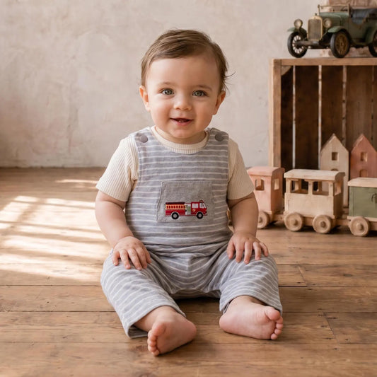 A baby sitting on a wooden floor wearing a gray and white striped overall romper with a red fire truck patch on the chest, in a warm, rustic room with toys in the background.