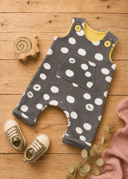 A baby's gray sleeveless romper with white polka dots, featuring a yellow inner collar and yellow button details, laid flat on a wooden surface with baby shoes, a wooden snail toy, and a pink blanket.