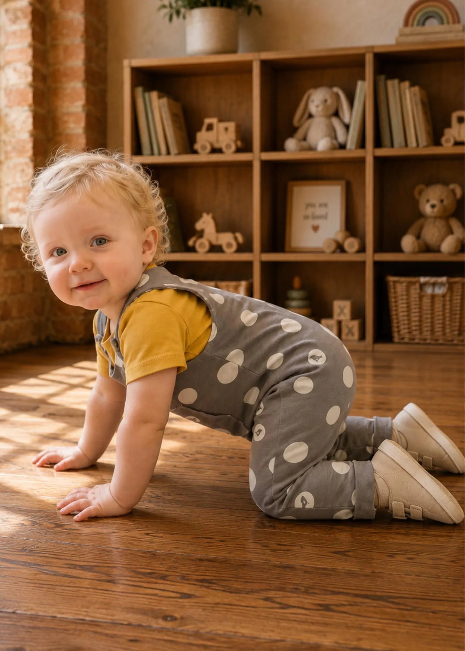 A toddler wearing a yellow short-sleeve shirt and gray polka-dot overall jumpsuit crawls on a wooden floor in a cozy room with wooden shelves and toys.