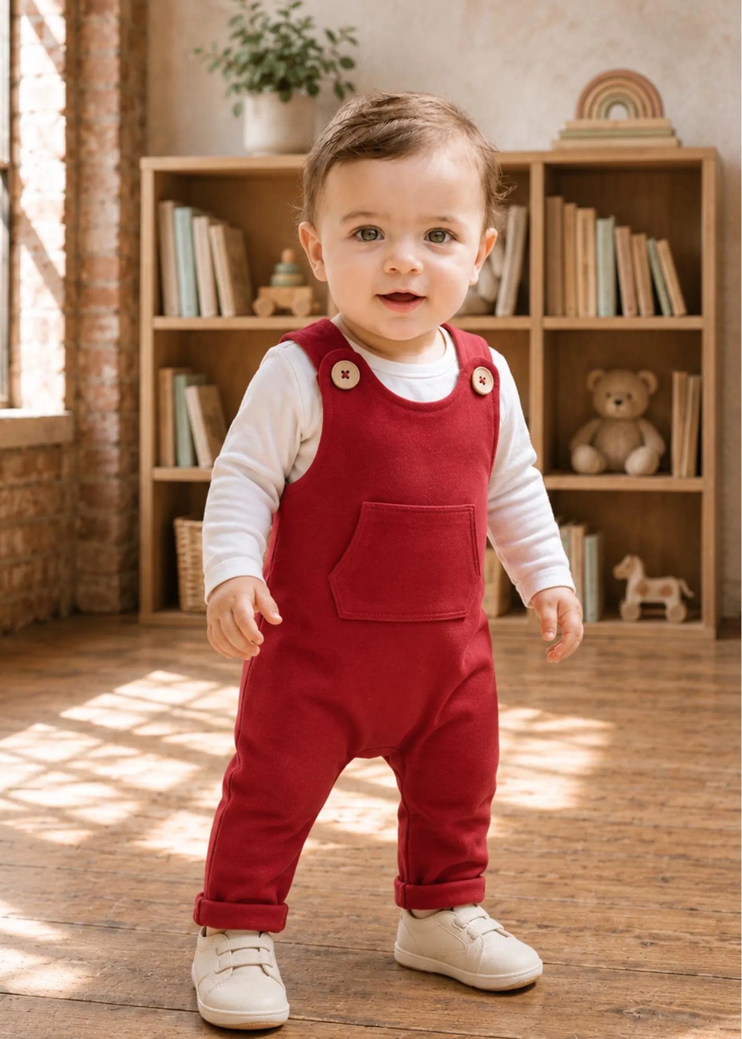 A toddler standing in a warm, sunlit room wearing red corduroy bib overalls over a white long-sleeve shirt and white shoes.