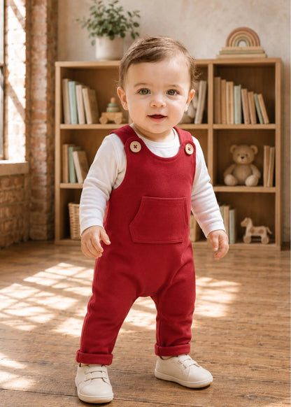 A toddler standing in a warm, sunlit room wearing red corduroy bib overalls over a white long-sleeve shirt and white shoes.