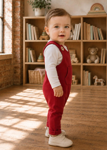 A toddler standing in a sunlit room with wooden floors and bookshelves, wearing red overall-style pants over a white long-sleeve shirt and white sneakers.