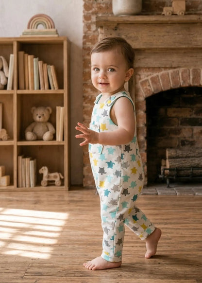 A toddler standing barefoot in a cozy room with a wooden floor and brick fireplace, wearing a sleeveless white jumpsuit covered in a colorful animal print of gray, yellow, and blue creatures.