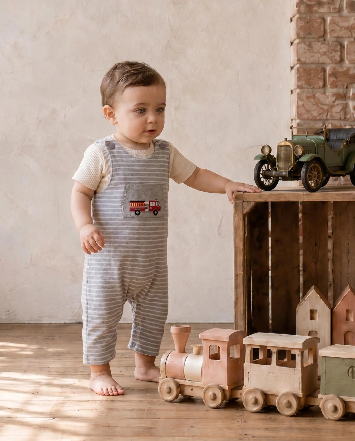A toddler stands barefoot in a rustic room, wearing a cream short-sleeve top and gray striped overall with a small red fire truck patch on the chest. The child is posed next to a wooden crate holding vintage toy cars, with a wooden train toy on the floor.