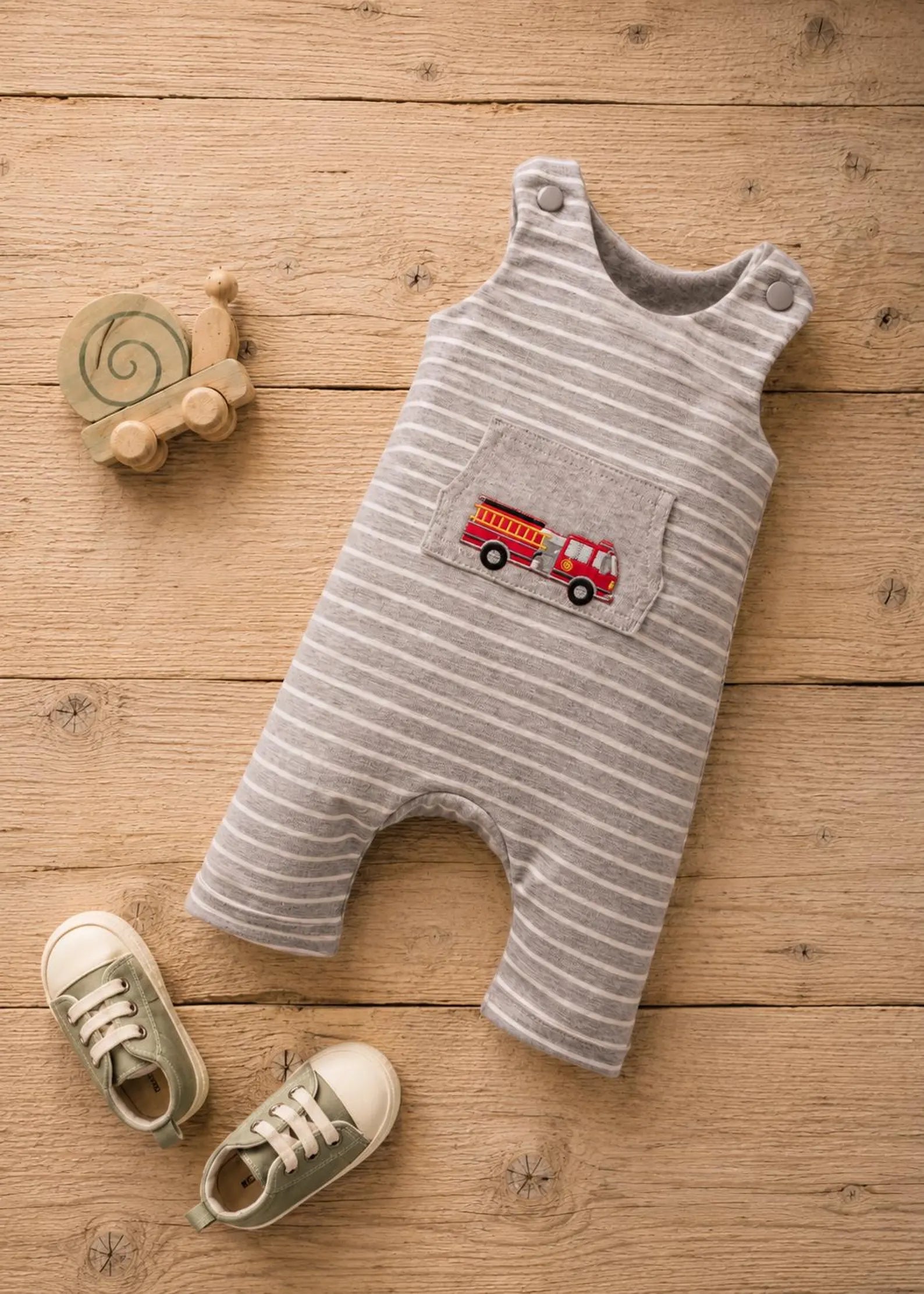 A baby romper laid flat on a wooden surface, featuring thin gray and white stripes and an embroidered red fire truck on the chest pocket. A wooden snail toy and a pair of baby shoes are styled around it.
