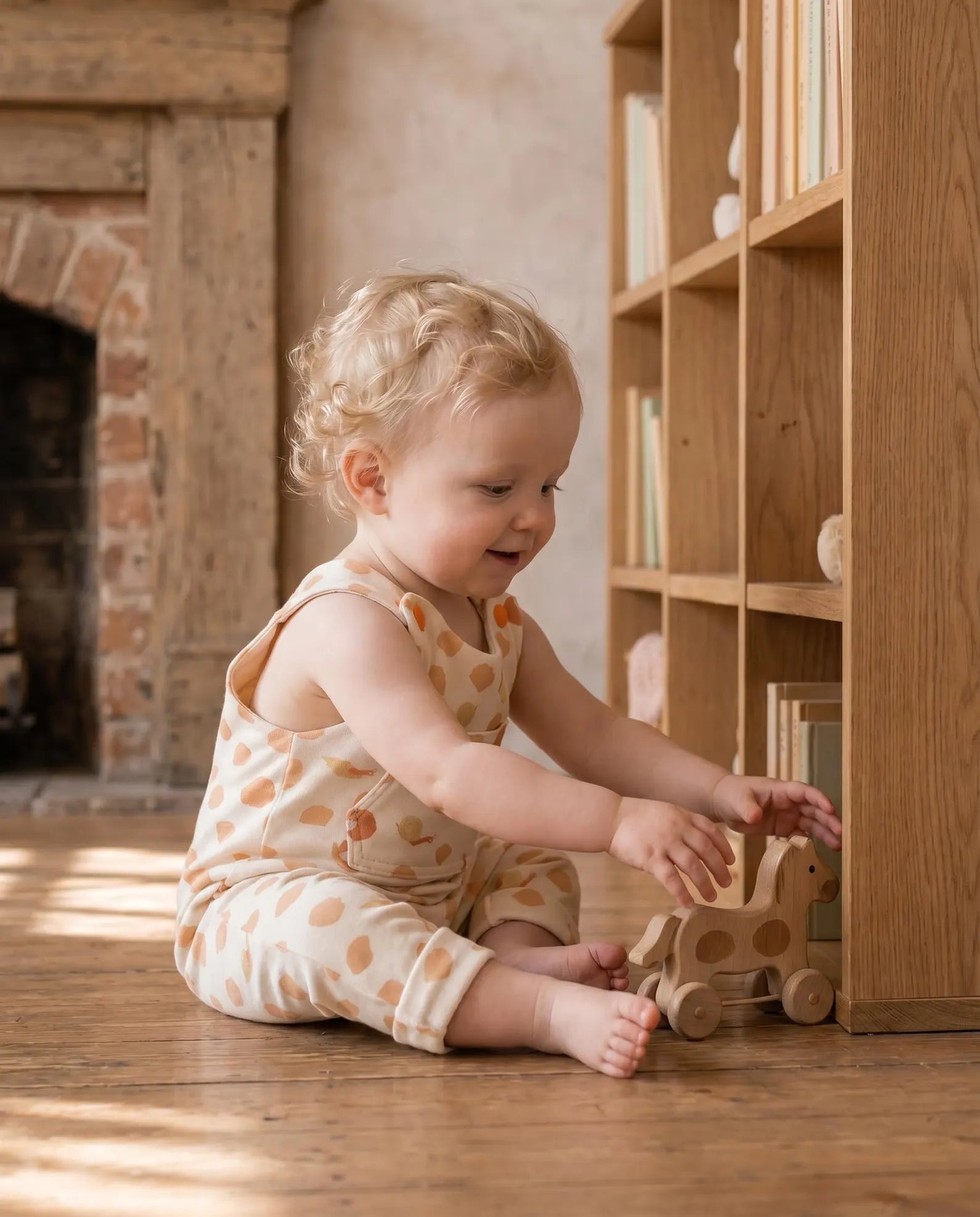 A toddler with curly blonde hair sits on a wooden floor in a cozy room, wearing a cream sleeveless romper with orange polka dots. The child is interacting with a wooden toy near a light wood bookshelf.