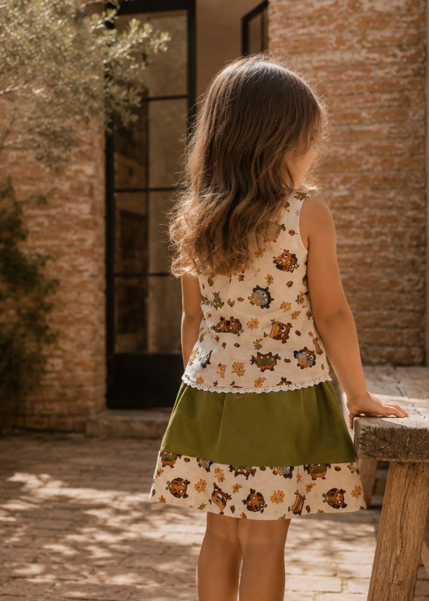A young girl standing outdoors with long brown hair wearing a sleeveless dress with a cream and brown owl print top, an olive green waistband, and a matching printed skirt. 