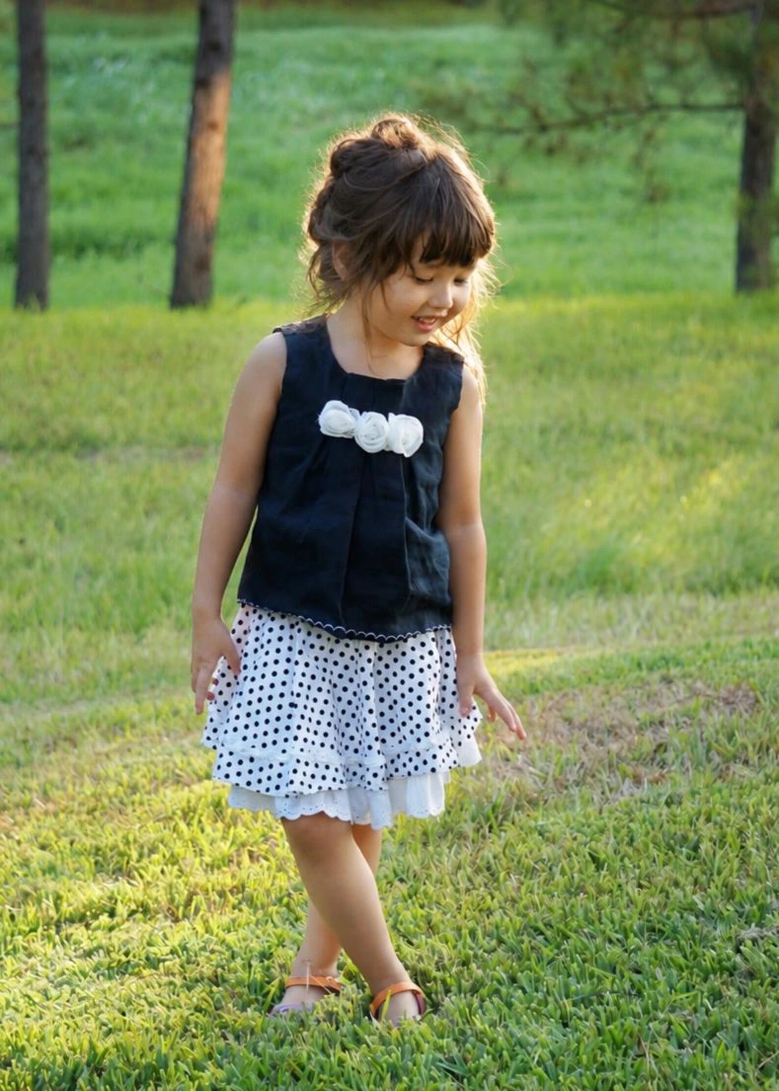 A young girl wearing a sleeveless black top with a white ruffle bow and a white skirt with black polka dots, standing on a grassy outdoor lawn.