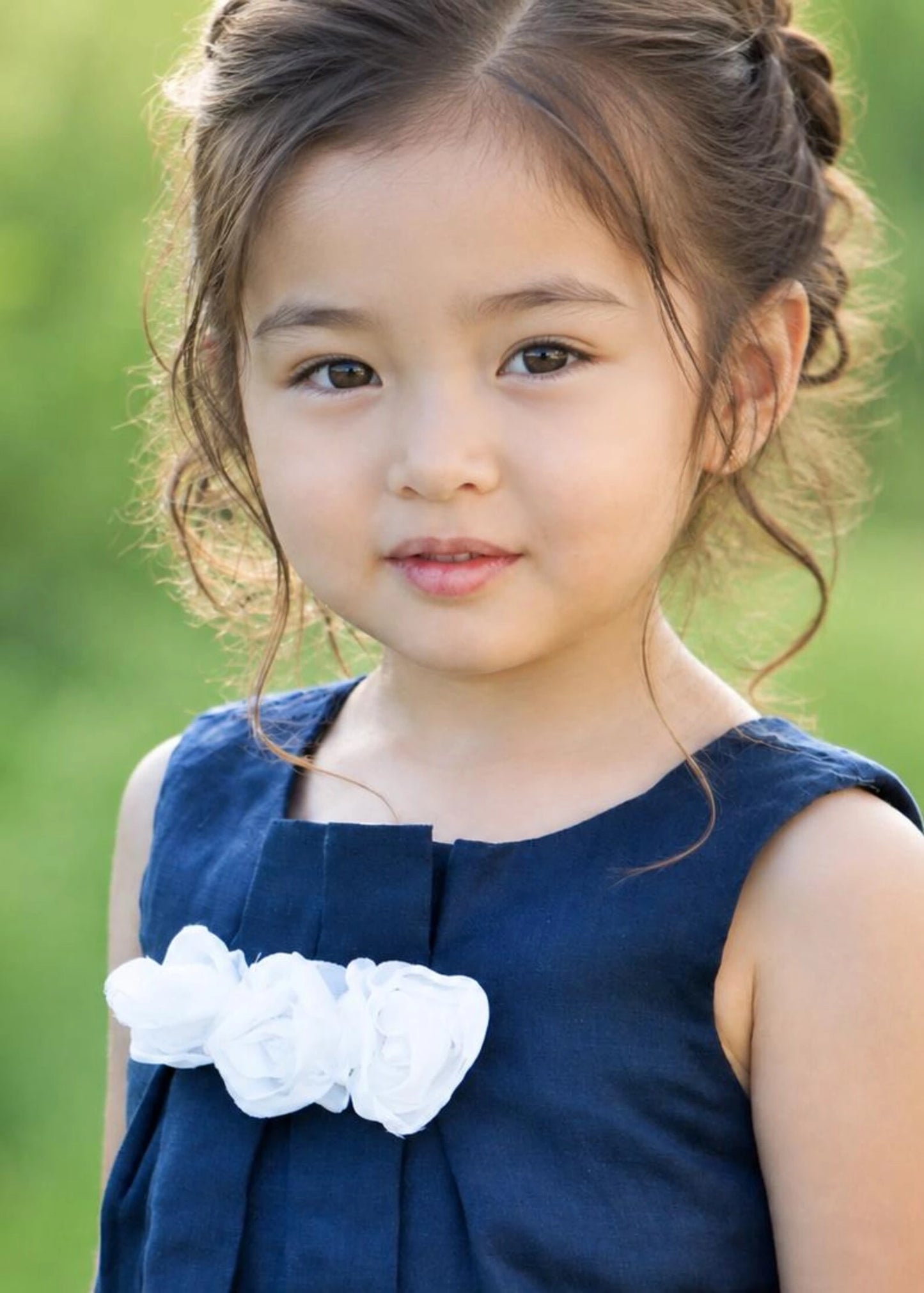 A young girl with brown hair styled in a half-up bun, wearing a navy blue sleeveless dress with a white fabric flower accent on the chest, set against a soft, blurred green background.