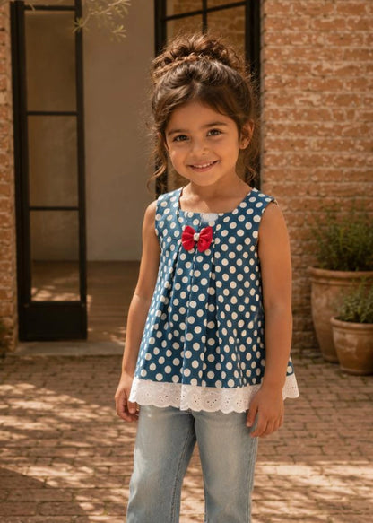 A young girl stands outdoors in front of a brick building, wearing a sleeveless blue and white polka dot top with a red bow and white lace hem, paired with light blue jeans.