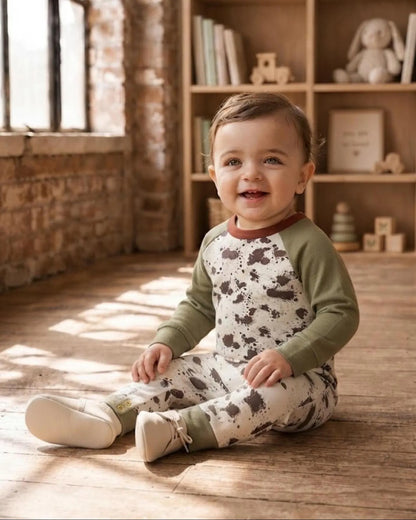 A baby sitting on a wooden floor in a sunlit room, wearing a one-piece raglan pajama with an olive green long-sleeve top and matching patterned pants.